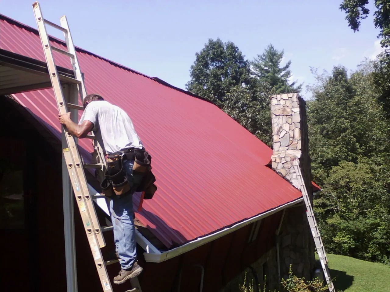 Roofer on a ladder beside a red metal roof and stone chimney — Soffit & Fascia Repair by Roofing Brothers in Woolford